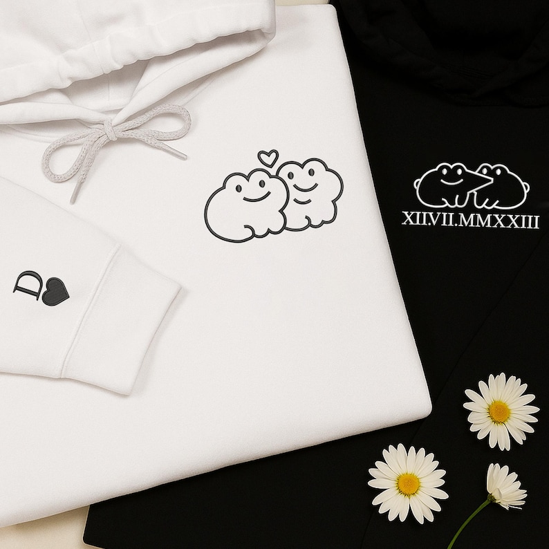 May include: White and black hoodies with matching designs. The white hoodie features a cloud design with two smiling faces and a heart. The black hoodie has a similar design with the Roman numerals XII.VII.MMXXIII. The white hoodie sleeve has a "D" and a heart.