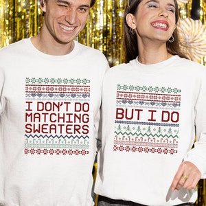 May include: Two white Christmas jumpers with a red and green patterned design. One jumper says "I don't do matching jumpers" and the other says "But I do".