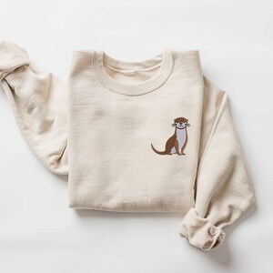 Peut inclure: Un sweat-shirt beige avec un graphique d'une loutre brune sur le devant. La loutre est assise et a un petit sourire sur son visage.