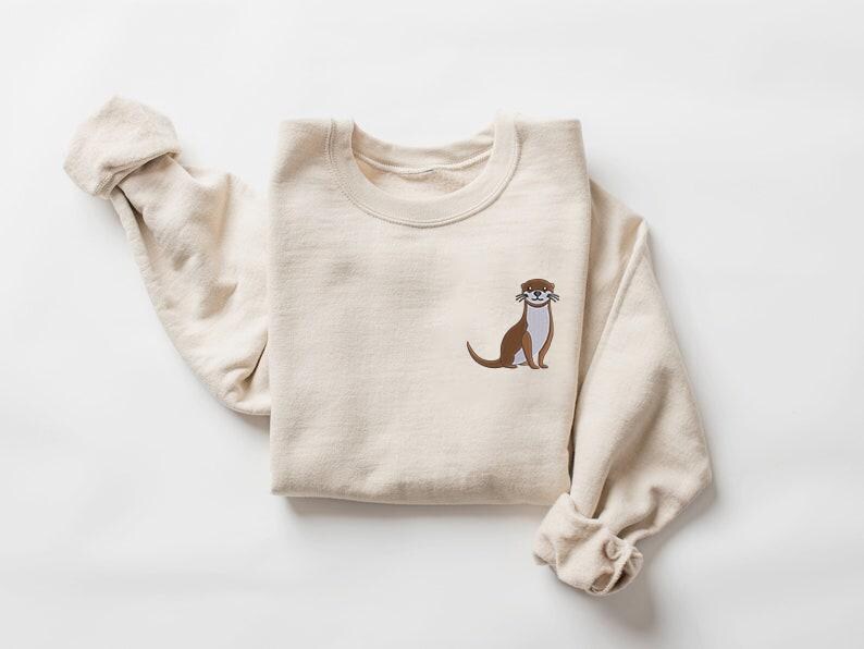 Peut inclure: Un sweat-shirt beige avec un graphique d'une loutre brune sur le devant. La loutre est assise et a un petit sourire sur son visage.