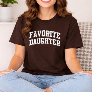 May include: A brown t-shirt with the words "FAVORITE DAUGHTER" printed in white, block letters. The shirt is a crew neck style and is being worn by a person.