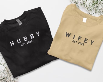 May include: Two folded t-shirts, one black with white text "HUBBY EST 2022" and one beige with black text "WIFEY EST 2022".