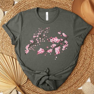 Puede incluir: Una camiseta verde oscuro con un dise&ntilde;o floral rosa y blanco. El dise&ntilde;o presenta una rama de cerezos en flor con flores rosas y centros blancos.