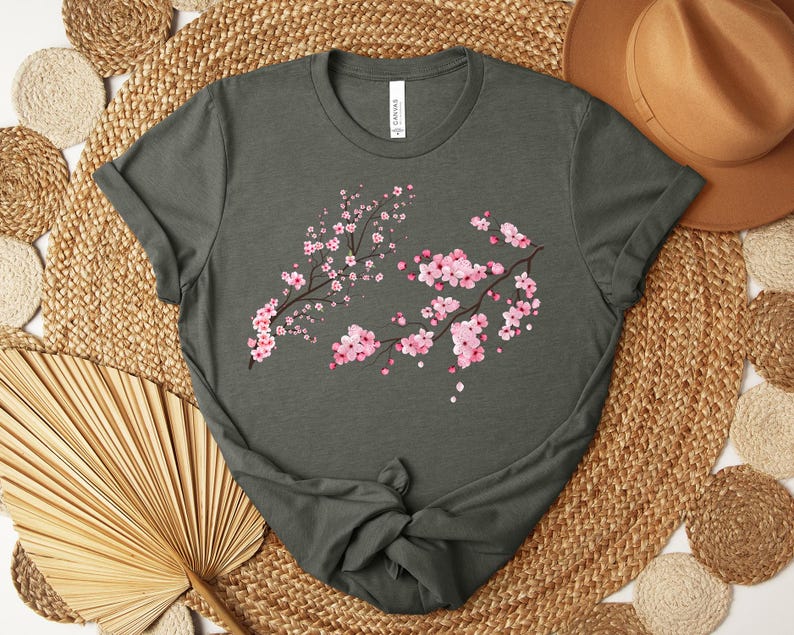 Puede incluir: Una camiseta verde oscuro con un dise&ntilde;o floral rosa y blanco. El dise&ntilde;o presenta una rama de cerezos en flor con flores rosas y centros blancos.