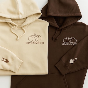 May include: Two hooded sweatshirts, one in beige and the other in brown, are shown. Each has an embroidered design of two cartoon frogs with a heart, and Roman numerals. The beige sweatshirt has an "H" and heart on the sleeve, the brown one has an "S" and heart.
