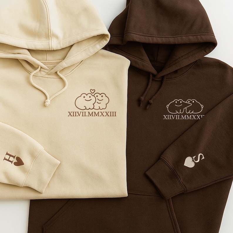 May include: Two hooded sweatshirts, one in beige and the other in brown, are shown. Each has an embroidered design of two cartoon frogs with a heart, and Roman numerals. The beige sweatshirt has an "H" and heart on the sleeve, the brown one has an "S" and heart.