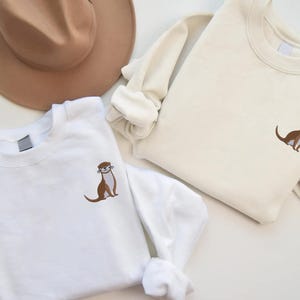 Peut inclure: Deux sweats &agrave; col rond blancs avec un motif de loutre brod&eacute;. La loutre est brune avec un ventre blanc et fait face &agrave; droite. Les sweats sont dispos&eacute;s sur une surface blanche.