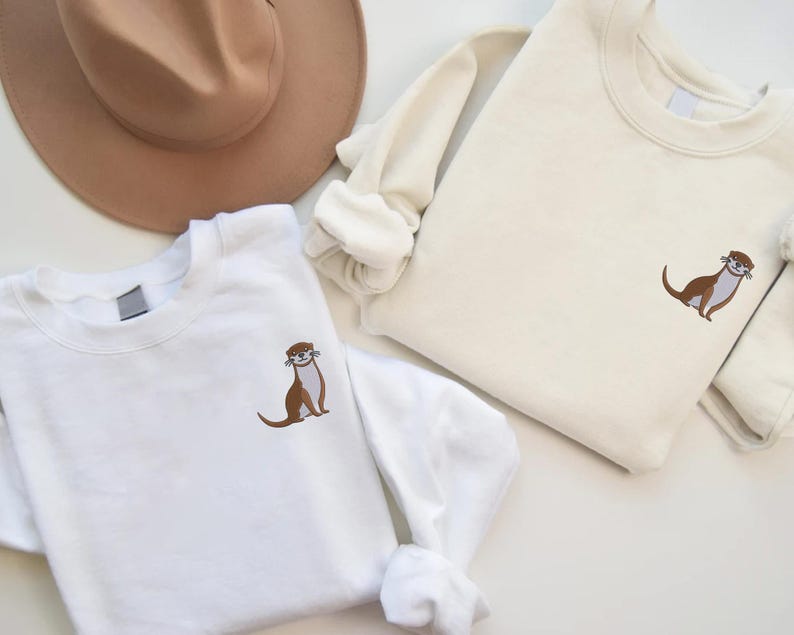 Peut inclure: Deux sweats &agrave; col rond blancs avec un motif de loutre brod&eacute;. La loutre est brune avec un ventre blanc et fait face &agrave; droite. Les sweats sont dispos&eacute;s sur une surface blanche.