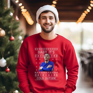 May include: A red Christmas sweater with a white snowflake pattern and the text "BABY IT'S COLD OUTSIDE". A man is pictured in the center of the sweater wearing a blue shirt.