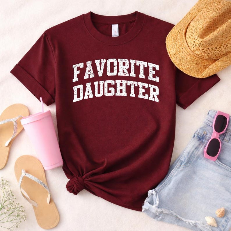 May include: A maroon t-shirt with the text "FAVORITE DAUGHTER" in white, distressed lettering. The shirt is styled with a straw hat, pink sunglasses, denim shorts, flip-flops, and a pink tumbler with a straw.