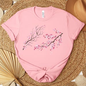 Puede incluir: Camiseta rosa con un gr&aacute;fico de dos ramas de cerezos en flor con flores rosas.