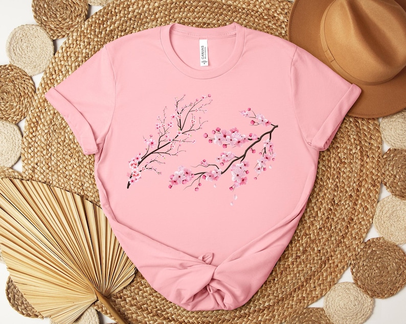 Puede incluir: Camiseta rosa con un gr&aacute;fico de dos ramas de cerezos en flor con flores rosas.