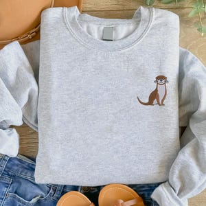Peut inclure: Un sweat-shirt gris avec une loutre brune brod&eacute;e sur le devant. La loutre est de face, la bouche ouverte et les pattes sur le sol.