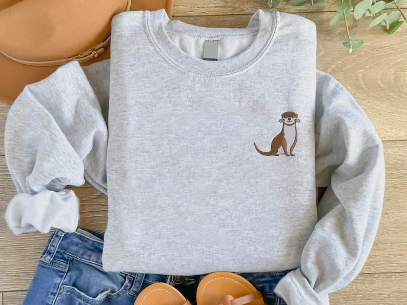 Peut inclure: Un sweat-shirt gris avec une loutre brune brod&eacute;e sur le devant. La loutre est de face, la bouche ouverte et les pattes sur le sol.