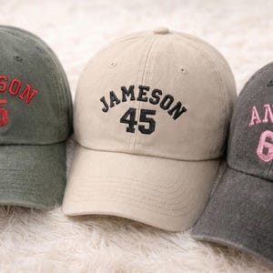 Peut inclure: Trois casquettes de baseball en vert olive, beige et gris anthracite. Chaque casquette présente un texte brodé : "GRAYSON 75" en rouge, "JAMESON 45" en noir et "ANSLIE 65" en rose. Les casquettes sont présentées sur une surface blanche et duveteuse.