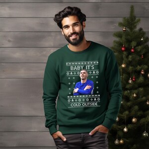 May include: A green Christmas sweater with a white snowflake pattern and the text "Baby it's cold outside". A man is wearing the sweater and a photo of a man is printed on the sweater.