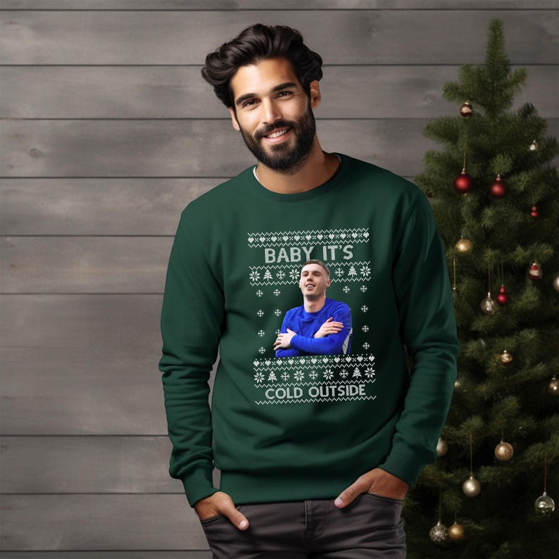 May include: A green Christmas sweater with a white snowflake pattern and the text "Baby it's cold outside". A man is wearing the sweater and a photo of a man is printed on the sweater.