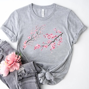 Puede incluir: Una camiseta gris con un dise&ntilde;o de flor de cerezo rosa. La camiseta est&aacute; hecha de un material suave y c&oacute;modo y tiene un corte holgado.