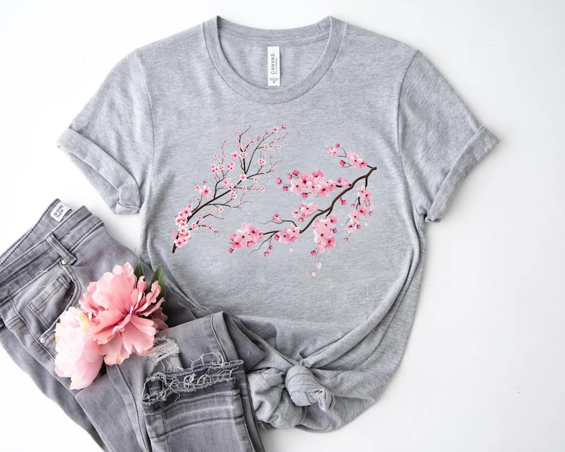 Puede incluir: Una camiseta gris con un dise&ntilde;o de flor de cerezo rosa. La camiseta est&aacute; hecha de un material suave y c&oacute;modo y tiene un corte holgado.