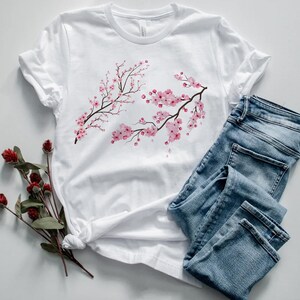 Puede incluir: Camiseta blanca con estampado de flores de cerezo rosas. La camiseta est&aacute; colocada sobre una superficie blanca con un par de pantalones vaqueros azules.