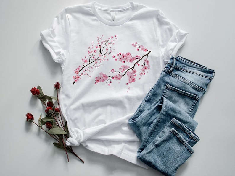 Puede incluir: Camiseta blanca con estampado de flores de cerezo rosas. La camiseta est&aacute; colocada sobre una superficie blanca con un par de pantalones vaqueros azules.