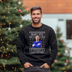 May include: A black Christmas sweater with a white snowflake pattern and a picture of a man in a blue shirt with the text "Baby It's Cold Outside".