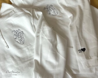 May include: Two white hoodies with black embroidery. One hoodie has a heart with a line drawing of a human heart. The other hoodie has a black heart with the letter "S" above it. The text "Customize - UK Store" is on the bottom of the image.