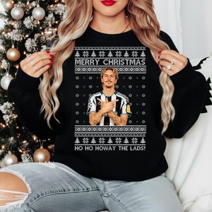 Puede incluir: Sudadera negra con un diseño festivo. La sudadera presenta el texto "MERRY CHRISTMAS" y "HO HO HOWAY THE LADS!" en blanco. Un gráfico de una persona con una camiseta a rayas blancas y negras está en el centro.