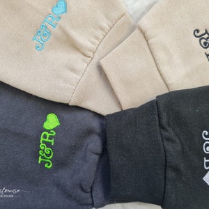 Pode incluir: Quatro sweatshirts de cores diferentes com o texto "J&R" ou "J&B" bordado nelas. Cada sweatshirt tem um pequeno cora&ccedil;&atilde;o bordado ao lado do texto. As sweatshirts s&atilde;o bege, preto, cinza e cinza escuro.