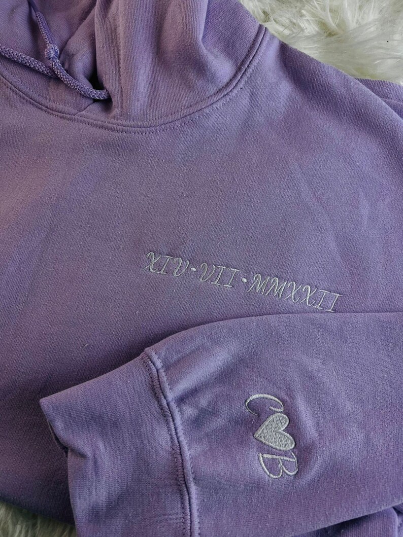 Peut inclure: Un sweat &agrave; capuche violet avec le texte "XIV - VII - MMXXII" brod&eacute; sur le devant et les lettres "C&B" avec un c&oelig;ur brod&eacute; sur la manche gauche.