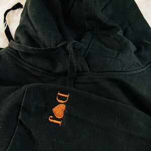 Peut inclure: Sweat-shirt &agrave; capuche noir avec un c&oelig;ur brod&eacute; orange et le texte "I Do It" sur le devant.