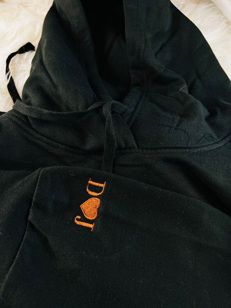 Peut inclure: Sweat-shirt &agrave; capuche noir avec un c&oelig;ur brod&eacute; orange et le texte "I Do It" sur le devant.