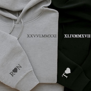 May include: Close-up of two hoodies, one gray and one black. The gray hoodie has the Roman numeral date XXV.VI.MMXXI printed on the chest and the initials "RON" with a heart on the sleeve. The black hoodie has the Roman numeral date XI.IV.MMXVII and the initials "T" with a heart.
