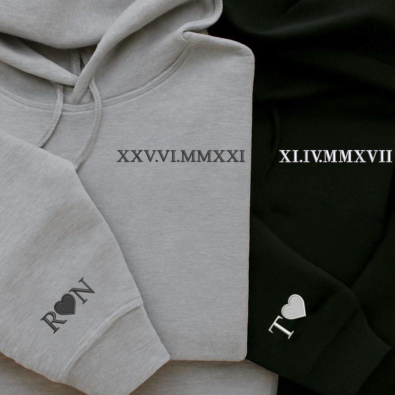 May include: Close-up of two hoodies, one gray and one black. The gray hoodie has the Roman numeral date XXV.VI.MMXXI printed on the chest and the initials "RON" with a heart on the sleeve. The black hoodie has the Roman numeral date XI.IV.MMXVII and the initials "T" with a heart.