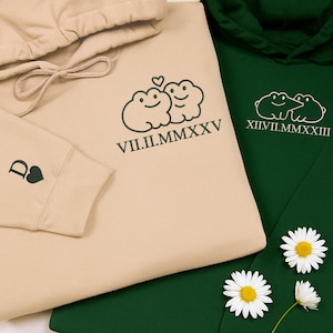May include: Two hoodies, one beige and one dark green, are displayed. The beige hoodie features a frog design with the Roman numeral date VII.II.MMXXV. The dark green hoodie has a similar frog design and the Roman numeral date XII.II.MMXXIII. Daisies are scattered around the hoodies.