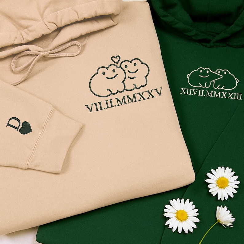 May include: Two hoodies, one beige and one dark green, are displayed. The beige hoodie features a frog design with the Roman numeral date VII.II.MMXXV. The dark green hoodie has a similar frog design and the Roman numeral date XII.II.MMXXIII. Daisies are scattered around the hoodies.