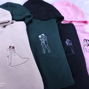 Pode incluir: Quatro sweatshirts com capuz em cores diferentes, cada uma com um design bordado diferente de duas pessoas se abra&ccedil;ando. Os designs incluem um casal em trajes de casamento, um casal em trajes de banho, um casal dan&ccedil;ando e um casal de m&atilde;os dadas. As sweatshirts est&atilde;o dispon&iacute;veis em bege, verde, preto e rosa.