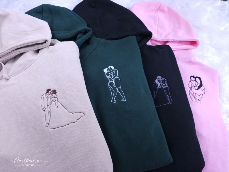 Pode incluir: Quatro sweatshirts com capuz em cores diferentes, cada uma com um design bordado diferente de duas pessoas se abra&ccedil;ando. Os designs incluem um casal em trajes de casamento, um casal em trajes de banho, um casal dan&ccedil;ando e um casal de m&atilde;os dadas. As sweatshirts est&atilde;o dispon&iacute;veis em bege, verde, preto e rosa.