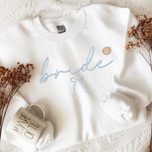 May include: A white sweatshirt embroidered with the word "bride" in light blue script. A small light blue bow is below the word. A small, round, wooden pin is on the upper right. A white mug with the words "MADE OF TE" is on the lower left.