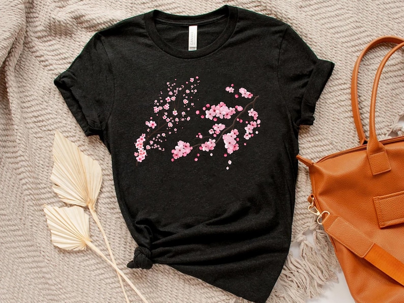 Puede incluir: Una camiseta negra con un dise&ntilde;o de flores de cerezo rosas. La camiseta est&aacute; hecha de un material suave y c&oacute;modo y tiene un corte holgado.