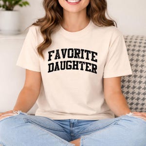 May include: A white ceramic mug with a black handle and the text "But First, Coffee" in a bold, black font. The mug is set against a blurred background, suggesting a cosy setting.