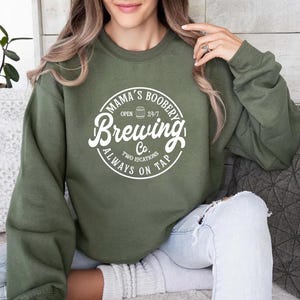 May include: A green sweatshirt with a white graphic that says "Mama's Boobery Brewing Co. Open 24/7 Two Locations Always On Tap".