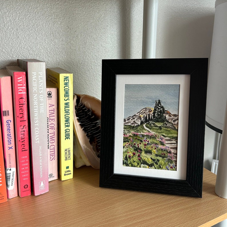 K&ouml;nnte beinhalten: Ein gerahmtes Aquarellgem&auml;lde einer Landschaft mit einem Weg, der einen H&uuml;gel hinauff&uuml;hrt, umgeben von B&auml;umen und Blumen. Das Gem&auml;lde befindet sich in einem schwarzen Rahmen und wird auf einer Holzoberfl&auml;che neben einer Reihe von B&uuml;chern ausgestellt.