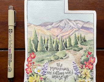 Könnte beinhalten: Aquarellmalerei der Utah-Landschaft in Form des Bundesstaates. Das Kunstwerk zeigt Berge, Bäume und Blumen mit dem Text "The mountains are calling and I must go." Ein Micron 005 Stift befindet sich neben dem Kunstwerk.