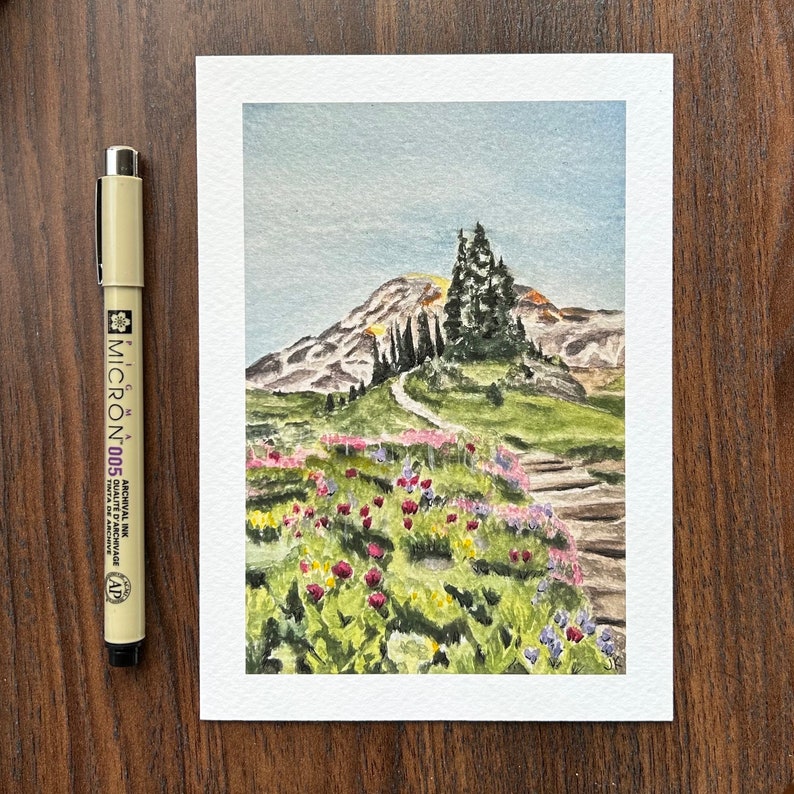 K&ouml;nnte beinhalten: Aquarellmalerei einer Berglandschaft mit einem Weg, der nach oben f&uuml;hrt. Das Gem&auml;lde zeigt einen Berg, B&auml;ume und bunte Blumen. Ein schwarzer und beiger Micron 005 Stift befindet sich neben dem Gem&auml;lde.
