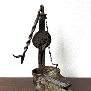 May include: An antique iron oil lamp featuring a rustic, weathered finish with visible oxidation. The lamp includes a vertical support rod, a circular backplate, a twisted metal handle, and a small metal chain connecting the reservoir to the frame.