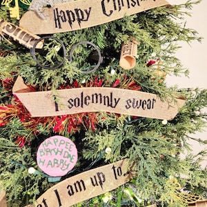 Può includere: Un albero di Natale decorato con vari ornamenti e striscioni. L'albero presenta una sciarpa, occhiali e striscioni con frasi come "Happy Christmas" e "I solemnly swear". Un ornamento rosa recita "Happee Birthdae Harry."