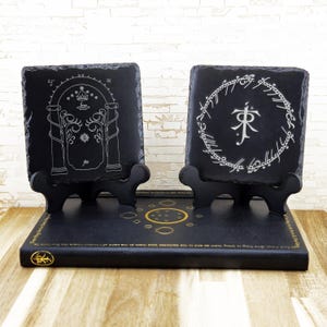 May include: Two black slate coasters with white etched designs, displayed on small black stands. One coaster features a door design, the other a circular inscription. The base has a gold emblem and text.