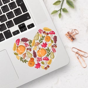 May include: A heart-shaped sticker with a fall theme. It features pumpkins, leaves, mushrooms, acorns, and berries in shades of orange, red, yellow, and brown.
