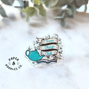 Puede incluir: Una tetera azul turquesa con asa y pico blancos, sentada sobre un libro abierto con una cubierta marrón. El libro está rodeado de diseños florales y una pancarta que dice "Finer Things Club".